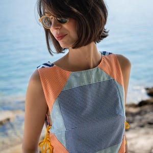Pode incluir: Um top cropped de patchwork com uma mistura de padrões e cores. O top apresenta vichy laranja, vichy azul e branco e um tecido azul claro com estampas de âncoras. Laços amarelos adornam as laterais. O top é sem mangas.