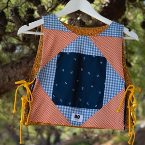 Può includere: Un gilet patchwork a tema nautico, con un quadrato blu navy con motivi di ancore bianche, circondato da tessuti a quadretti arancioni e quadretti blu. Il gilet ha lacci gialli e le lettere "DO" su una piccola etichetta.