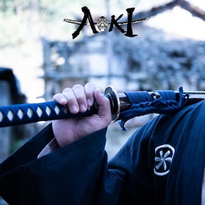 May include: A close-up of a katana sword with a blue handle and a black blade. The sword is held by a person wearing a dark blue kimono. The word "Aoki" is written in Japanese characters above the sword.