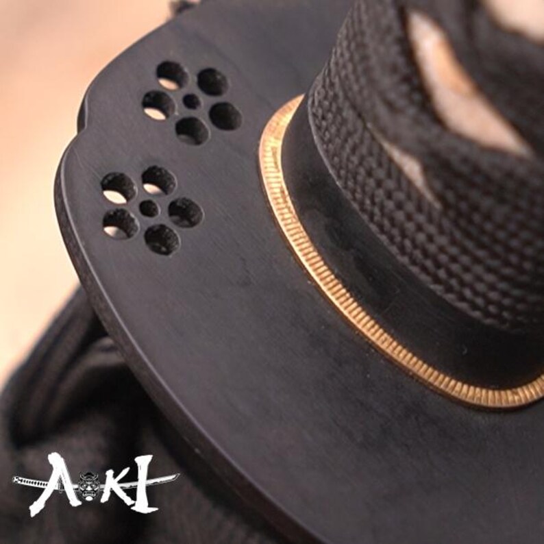 May include: Close-up of a black sword handle with a gold trim and a decorative design with small holes. The handle is made of wood and has a textured surface.