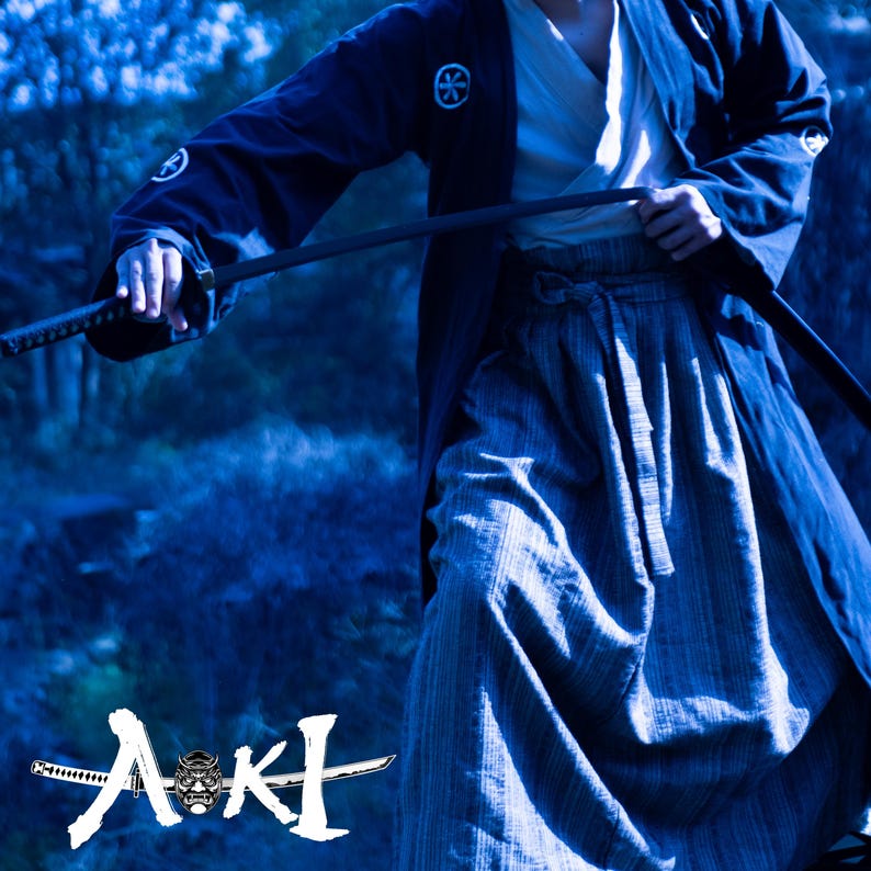 May include: A person wearing a dark blue kimono with a white symbol on the chest, holding a black katana sword. The person is standing in a forest setting with a blue and green color scheme. The word "AKI" is written in white with a stylized demon mask in the center.