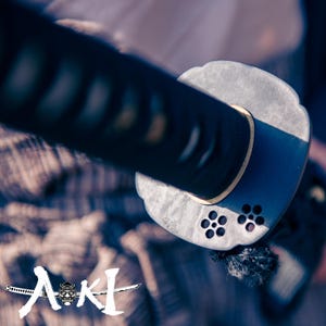 May include: Close-up of the black metal handle of a katana sword. The handle is decorated with a silver ring and a pattern of small circles. The sword is resting on a fabric surface.