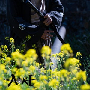 May include: A person wearing a black kimono with a sword strapped to their side, standing in a field of yellow flowers. The text "AKI" is visible in the lower center of the image.