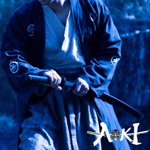 May include: A person wearing a blue kimono and holding a katana sword. The sword is sharp and has a black handle. The person is standing in front of a dark blue background. The text "AKI" is visible in the lower right corner of the image.