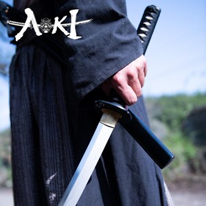 May include: A person wearing a black robe holds a silver katana sword with a black scabbard. The sword is partially drawn from the scabbard. The word "AKI" is written in white on the robe.