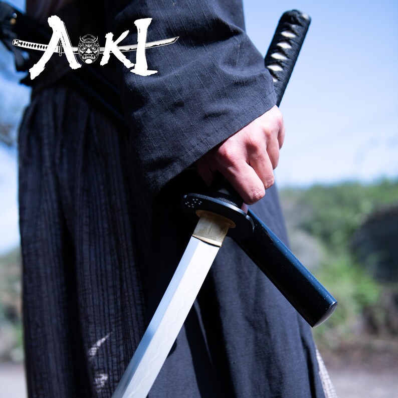May include: A person wearing a black robe holds a silver katana sword with a black scabbard. The sword is partially drawn from the scabbard. The word "AKI" is written in white on the robe.