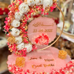May include: A pink and gold decorative piece with white roses, pink and gold accents, and the date 22-09-2025. The piece includes the names Zain & Mina and the words 'Baat Pakki'.
