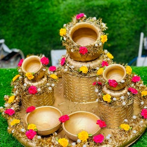 May include: A gold decorative tray featuring small pots and bowls, decorated with yellow and pink flowers and white pearls. The tray and vessels have a textured, woven design, creating an elegant and festive display, suitable for a special occasion.