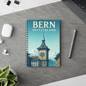 May include: A spiral-bound notebook featuring a stylized illustration of Bern, Switzerland. The cover displays the city's name and a clock tower against a backdrop of mountains and a blue sky. The notebook is on a gray surface.