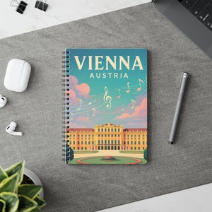 May include: A notebook with the words "VIENNA AUSTRIA" on the cover. The cover shows a building illustration, musical notes, and a sky with clouds. A pen, earbuds, and a laptop are also visible. The notebook is spiral-bound.