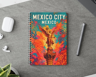 May include: Spiral-bound notebook with a colourful illustration of the Angel of Independence in Mexico City. The cover reads "MEXICO CITY MEXICO" in white, set against a background of orange, red, and blue. A pen and other items are visible.