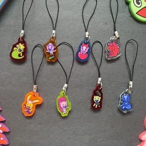 May include: A collection of colorful cartoon character charms with black lanyards. The charms feature various designs, including a green lizard, a purple bunny, and a red hedgehog. The charms are attached to silver-colored metal clasps.