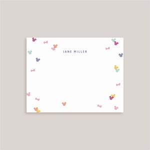 May include: A white rectangular stationery card featuring the centered text JANE MILLER in dark blue. The card is decorated with a scattered pattern of small, colorful mouse-ear silhouettes and tiny bows in shades of pink, yellow, blue, and green.