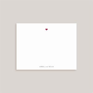May include: A rectangular white stationery card featuring a small, centered dark red heart icon near the top edge. The bottom of the card displays the text "APRIL and RYAN" in a clean, dark gray serif font, centered horizontally on the white surface.