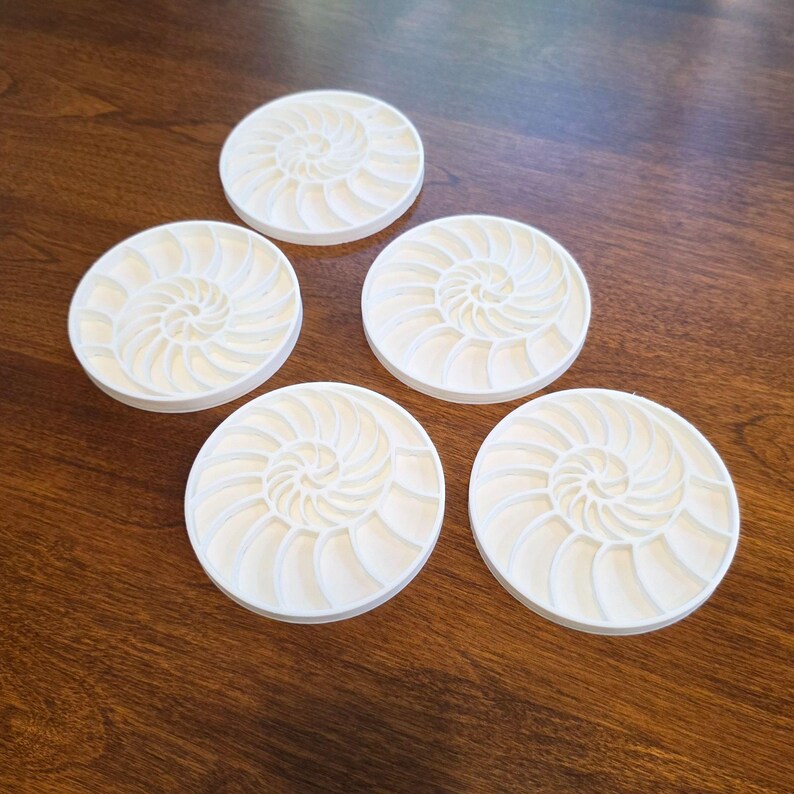 Puede incluir: Cinco posavasos redondos de color blanquecino con un dise&ntilde;o de concha de nautilus. Los posavasos est&aacute;n dispuestos sobre una superficie de madera marr&oacute;n oscuro. Cada posavasos presenta un patr&oacute;n en espiral.