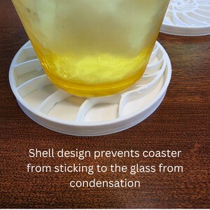 Puede incluir: Un posavasos blanco con forma de concha con un vaso de l&iacute;quido amarillo encima. El dise&ntilde;o del posavasos evita que se pegue por la condensaci&oacute;n. Texto: El dise&ntilde;o de concha evita que el posavasos se pegue al vaso.