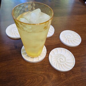 Puede incluir: Un vaso alto, te&ntilde;ido de amarillo, lleno de hielo y un l&iacute;quido amarillo, descansa sobre un posavasos blanco con un dise&ntilde;o de concha de nautilus. Varios posavasos a juego est&aacute;n dispersos.