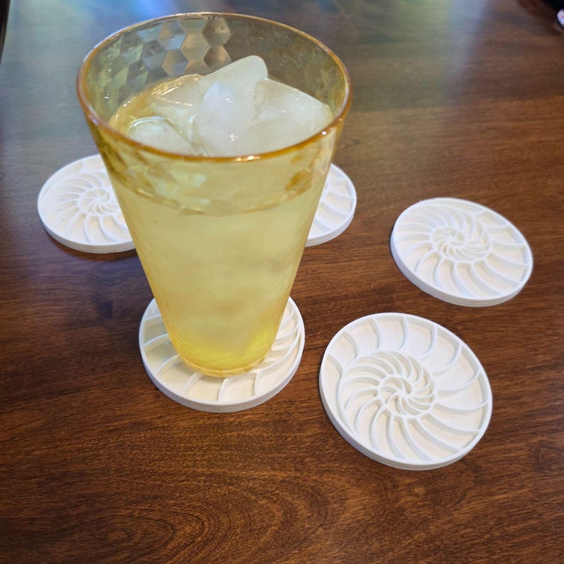 Puede incluir: Un vaso alto, te&ntilde;ido de amarillo, lleno de hielo y un l&iacute;quido amarillo, descansa sobre un posavasos blanco con un dise&ntilde;o de concha de nautilus. Varios posavasos a juego est&aacute;n dispersos.