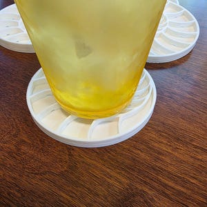 Puede incluir: Un vaso transparente con l&iacute;quido amarillo descansa sobre un posavasos blanco con forma de nautilus. El posavasos est&aacute; hecho de una serie de l&iacute;neas curvas y segmentadas. El posavasos est&aacute; sobre una superficie de madera marr&oacute;n oscuro.