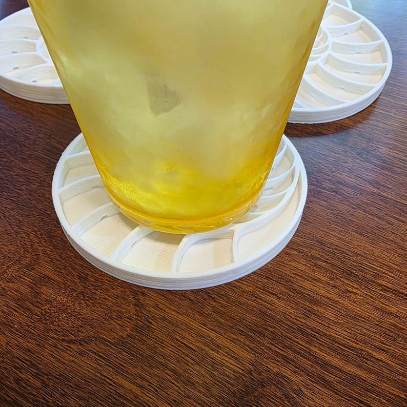 Puede incluir: Un vaso transparente con l&iacute;quido amarillo descansa sobre un posavasos blanco con forma de nautilus. El posavasos est&aacute; hecho de una serie de l&iacute;neas curvas y segmentadas. El posavasos est&aacute; sobre una superficie de madera marr&oacute;n oscuro.