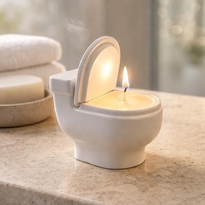 Könnte beinhalten: Eine weiße, keramikförmige Toilettenkerze mit leicht geöffnetem Deckel und einem brennenden Docht. Die Kerze ist mit gelbem Wachs gefüllt. Der Hintergrund umfasst einen Stapel weißer Handtücher und ein Stück Seife in einer kleinen Schale.