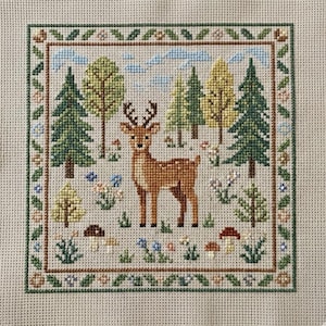 May include: Cross-stitch embroidery depicting a deer in a woodland setting. The deer is brown with white spots, surrounded by green and yellow trees, blue flowers, and mushrooms. The design is framed with a green and brown floral border.