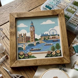 Könnte beinhalten: Ein gerahmtes Kreuzstichbild, das eine Londoner Stadtlandschaft darstellt, einschließlich Big Ben, dem London Eye und der Themse. Das Kunstwerk befindet sich in einem Holzrahmen. Die Farbpalette umfasst Blau-, Grün-, Braun- und Beigetöne.