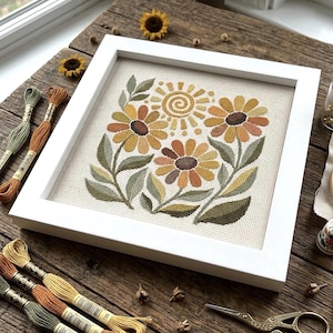 May include: A square white frame containing a cross-stitch embroidery of three sunflowers and a stylized sun. The design features shades of yellow, orange, and olive green on a light fabric. Several skeins of embroidery floss are arranged nearby.
