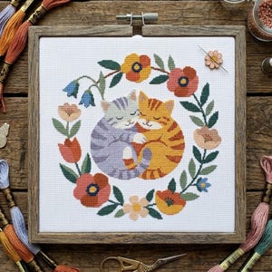 May include: Cross-stitch embroidery of two hugging cats, one gray and one orange, surrounded by a floral wreath. The artwork is framed in a square wooden frame. The background is a wooden surface with embroidery floss and tools.