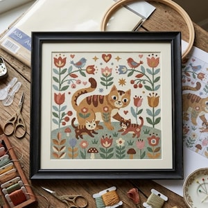 May include: A framed cross-stitch picture depicting a cat family in a garden setting. The design features a mother cat and two kittens, surrounded by flowers, birds, and mushrooms. The colour palette includes orange, brown, and green.