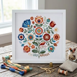 May include: A square white frame containing a cross-stitch embroidery of a symmetrical floral arrangement. The design features stylized flowers in shades of red, orange, blue, and green on a white fabric background, centered within the frame.