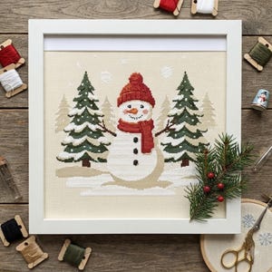 May include: A framed cross-stitch embroidery featuring a central snowman wearing a red knit hat and scarf, flanked by two green pine trees with white snow accents. The design is set against a light beige fabric background with white snowflakes.