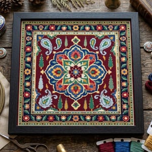 May include: A square cross-stitch embroidery piece framed in black, featuring a central floral medallion in blue, green, red, and yellow on a deep burgundy background, surrounded by paisley motifs and a decorative border of small flowers.