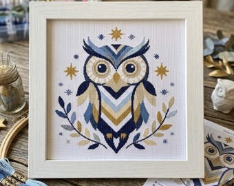 May include: A framed cross-stitch embroidery featuring a stylized owl in shades of navy blue, light blue, and gold. The owl is centered, surrounded by small gold stars and blue leaf sprigs, all stitched onto a white grid fabric within a white frame.
