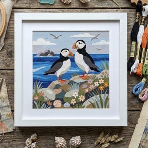 May include: A framed cross-stitch embroidery featuring two puffins standing on a rocky coastal cliff with blue ocean water, distant islands, and small white flowers in the foreground. The artwork is enclosed in a square white frame.
