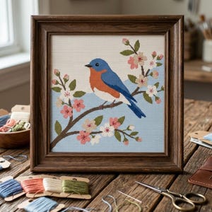 May include: Framed needlepoint art featuring a bluebird perched on a flowering branch. The bird has blue, orange, and white feathers. The artwork is set against a light blue background and framed in a dark wood. The scene includes pink and white blossoms.