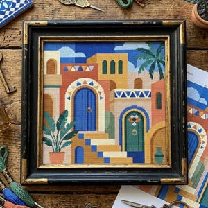 May include: A square cross-stitch embroidery depicting a Mediterranean-style architectural scene with yellow, orange, and beige buildings, a bright blue arched door, a green door, a palm tree, and a potted plant, framed in a black and gold frame.