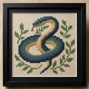 Peut inclure: Broderie au point de croix d'un serpent enroulé avec un corps jaune et bleu sarcelle, une langue rouge et des yeux verts, entouré de feuilles vertes. L'œuvre est encadrée dans un cadre carré noir.