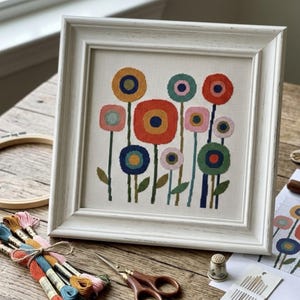 May include: A framed cross-stitch embroidery featuring a pattern of stylised flowers in shades of orange, blue, green, and pink. The artwork is displayed in a white distressed wooden frame, accompanied by embroidery floss, scissors, and needles.