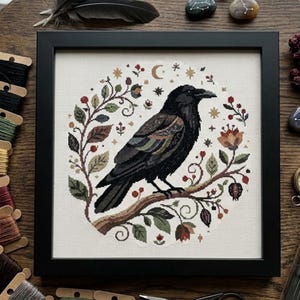 May include: A framed cross-stitch embroidery featuring a black raven perched on a branch, surrounded by a circular wreath of leaves, red berries, small flowers, a crescent moon, and stars, all stitched on a light beige fabric background.