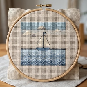 Può includere: Ricamo a punto croce di una barca a vela sull'acqua, incorniciato in un cerchio di legno. Il disegno presenta una barca blu, acqua blu, un cielo azzurro e soffici nuvole bianche. Il tessuto è di colore beige neutro.