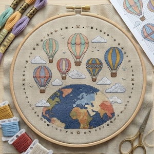 May include: A cross-stitch embroidery hoop featuring a colorful design of hot air balloons floating above a detailed depiction of the Earth. The design includes clouds and small birds, with embroidery floss and scissors nearby.