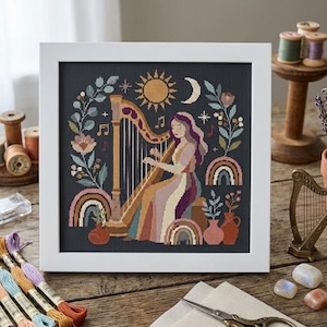 May include: A framed cross-stitch embroidery featuring a person playing a harp, surrounded by floral motifs, a sun, a crescent moon, rainbows, and musical notes on a dark background, all enclosed within a simple white square frame.