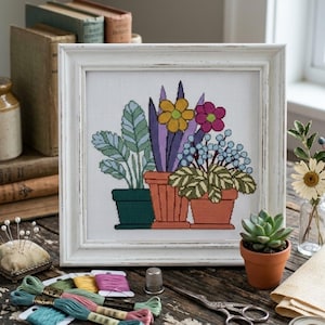 May include: A framed cross-stitch embroidery featuring three potted plants with green, yellow, purple, and pink foliage and flowers. The design is displayed in a white wooden frame, surrounded by embroidery floss, a thimble, and scissors.