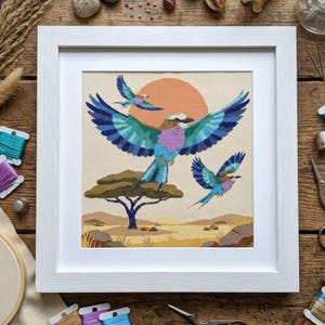 Könnte beinhalten: Eine gerahmte Kreuzstichstickerei mit drei bunten Vögeln im Flug vor einem beigen Hintergrund mit einer großen orangefarbenen Sonne, einem flachen Akazienbaum und einer Wüstenlandschaft. Das Kunstwerk befindet sich in einem quadratischen weißen Holzrahmen.