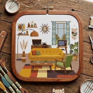 May include: A square wooden embroidery hoop containing a cross-stitch pattern of a living room interior. The design features a yellow sofa, a green armchair, a coffee table, a floor lamp, a sunburst clock, a television, and a potted plant.
