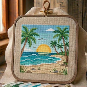 May include: Cross-stitch embroidery depicting a tropical beach scene. The artwork shows palm trees, a setting sun, and ocean waves in shades of blue, green, and yellow. It is framed in a square wooden hoop with a canvas backing.
