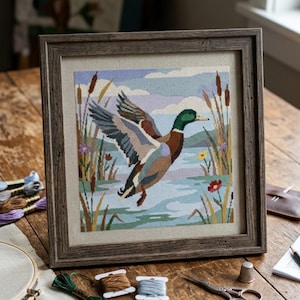 May include: Framed cross-stitch artwork of a mallard duck in flight over a lake, surrounded by reeds and flowers. The duck has green, brown, and blue plumage. The frame is a rustic, dark wood. The scene is set against a light blue sky.