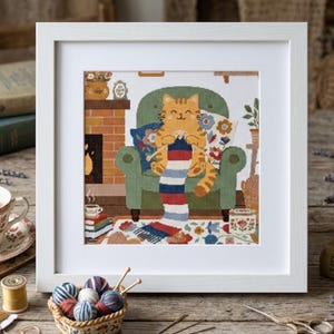 May include: A framed cross-stitch embroidery depicting an orange tabby cat sitting in a green armchair, knitting a long striped scarf. The scene includes a brick fireplace, a stack of books, and a floral rug, all encased in a white frame.