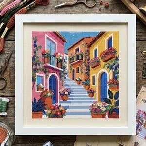 May include: A framed cross-stitch embroidery depicting a Mediterranean street scene with pink and yellow buildings, blue arched doors, and a central stone staircase. The artwork features potted flowers, climbing vines, and balconies in a vibrant palette.